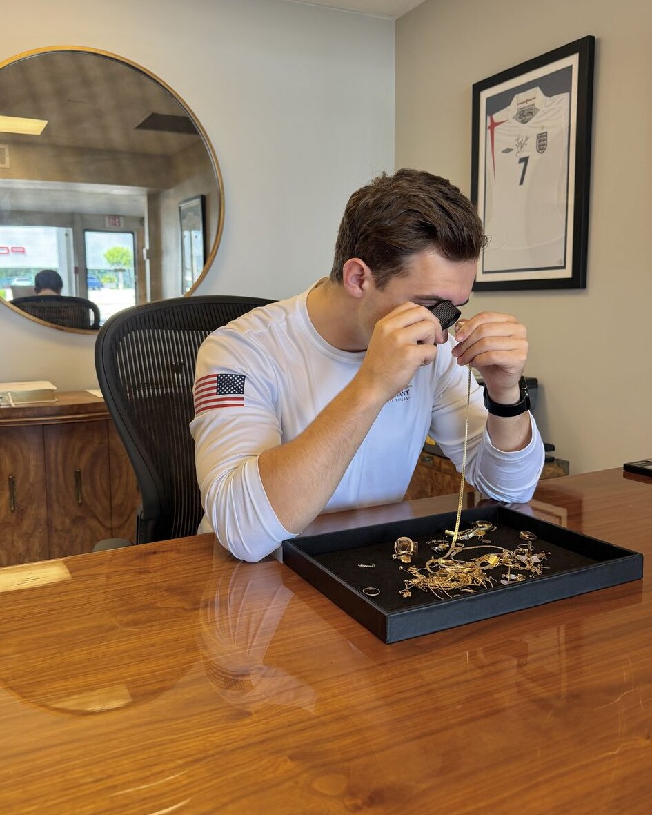 A Covenant evaluator examining a gold chain through a jeweler's loupe, with a tray of gold jewelry on the desk